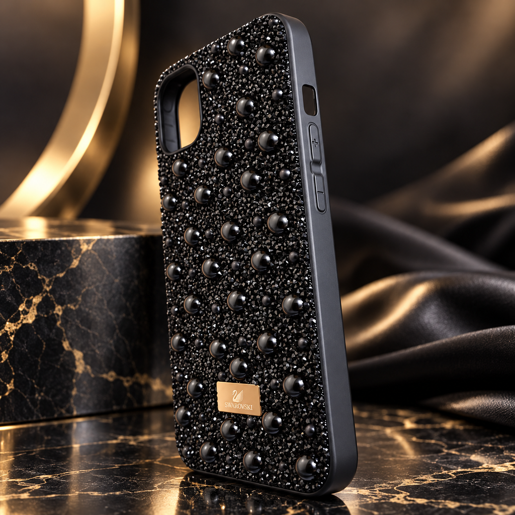 A black phone case covered in black rhinestones and raised rhinestone studs, with a small gold brand plaque reading 'SWAROVSKI'. The case is shown standing on a dark marble surface with draped black fabric and a gold-toned decorative object in the background.