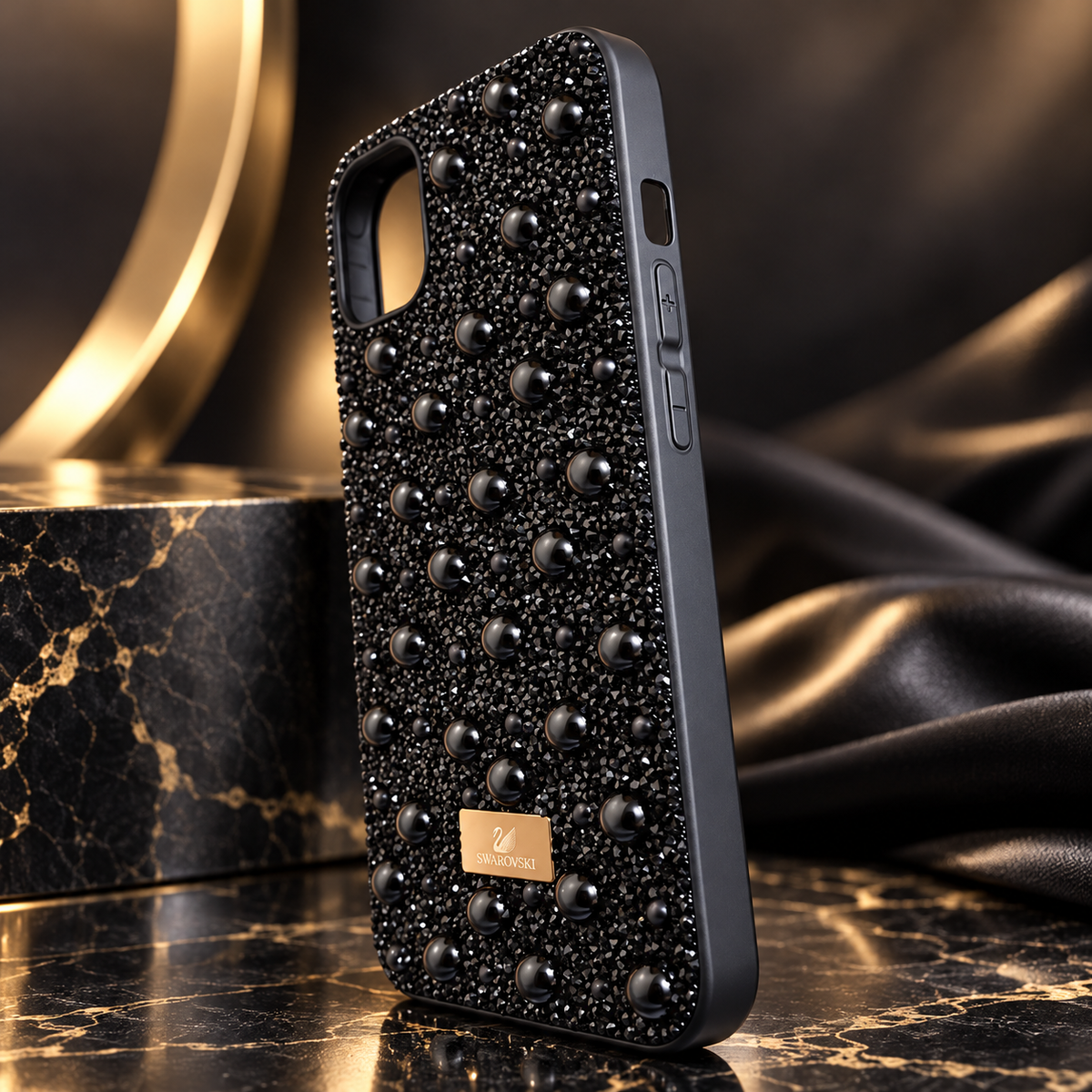 A black phone case covered in black rhinestones and raised rhinestone studs, with a small gold brand plaque reading 'SWAROVSKI'. The case is shown standing on a dark marble surface with draped black fabric and a gold-toned decorative object in the background.