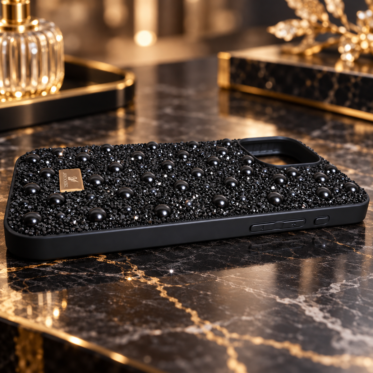 A black phone case covered in black pearls and beads, resting on a black and gold marble surface.