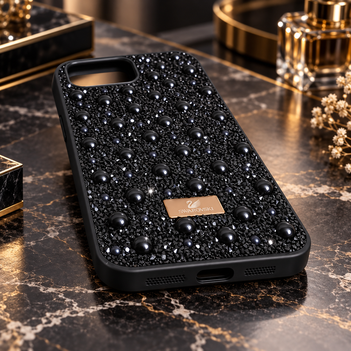 A black phone case decorated with black pearls and rhinestones, featuring a gold metal logo plate that reads 'SWAROVSKI'. The case is displayed on a dark marble surface with luxury accessories in the background.