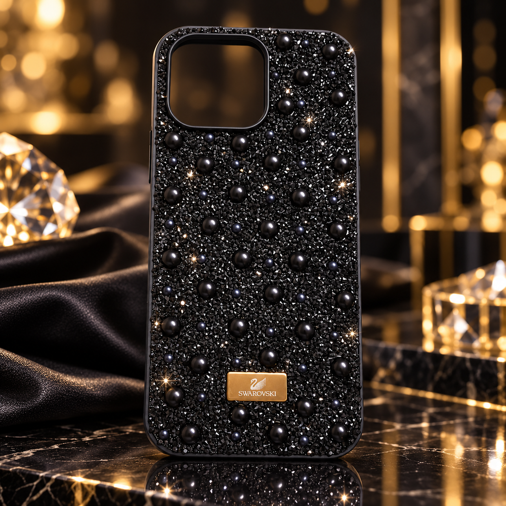 A black rhinestone & pearl phone case embellished with raised Swarovski crystal beads and a gold brand plaque, displayed on a dark marble surface with luxury props.