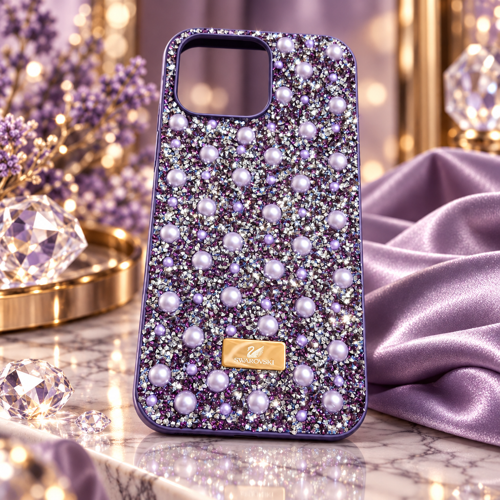 A purple rhinestone 16 pro max phone case decorated with faux pearls and a gold-tone Swarovski logo plaque, displayed on a marble surface with purple and crystal props.