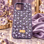 A purple rhinestone 16 pro max phone case decorated with faux pearls and a gold-tone Swarovski logo plaque, displayed on a marble surface with purple and crystal props.