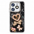 A clear phone case filled with black glitter and decorated with three decorative brooch designs: a heart-shaped brooch with pearls and a gold interlocking CC logo, a pearl-and-gold bow, and a pink-and-gold heart ribbon brooch. .